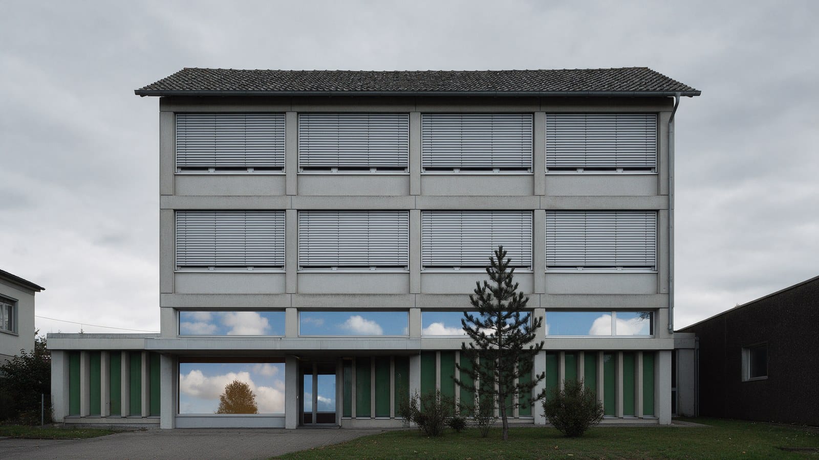 Frontalaufnahme eines schlichten dreigeschossigen Verwaltungsgebäudes bei bedecktem Himmel, sämtliche Storen der oberen Geschosse sind heruntergelassen, in den Fenstern des Erdgeschosses spiegeln sich Wolken