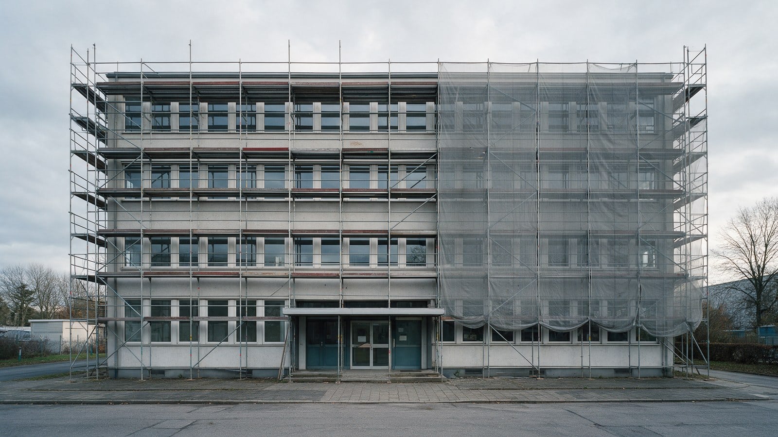 Vierstöckiges Bürogebäude bei bedecktem Himmel, die ganze Fassade eingerüstet, die rechte Hälfte mit grauem Baunetz verhüllt, der Eingang in der Mitte bleibt offen