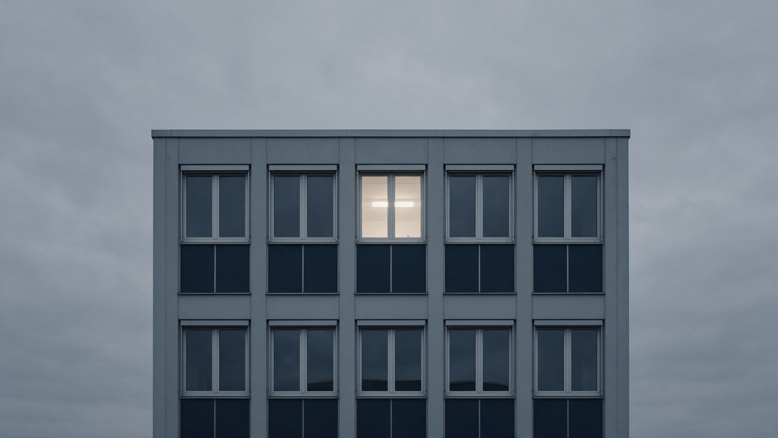 Fassade eines grauen Bürogebäudes vor bedecktem Himmel, fünf gleiche Fenster pro Reihe, nur in einem Fenster der oberen Reihe brennt warmes Licht